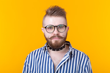 Young positive trendy man hipster with a mustache beard and fetish necklace in shirt posing on a yellow background. Concept of rock and subculture.