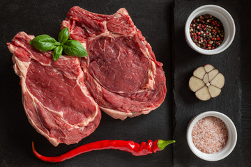 Raw fresh Beef steak with bone, spices and seasonings on stone cutting board. Top view, close upon black background