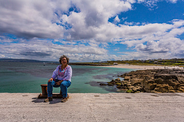 Mexican woman sitting on the shore of the pier of Inis Oirr Island with the beach, limestone rocks and the village in the background, sunny day in Inisheer, island in the west of the coast of Ireland