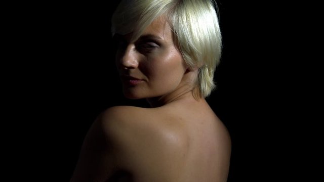 Naked blonde girl is posing sideways in the camera. Black background zoom out shot.