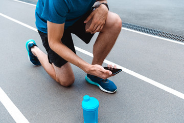 cropped view of young sportsman on running track with blue sports bottle and smartphone