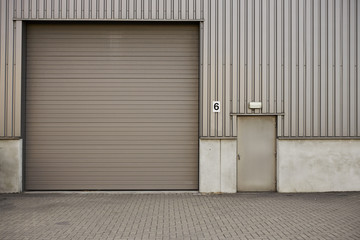 Outdoor shot of gray metal roll-down shutters on vertical profiled sheeting of industrial building. Garage for vehicles with locked door, clean paved surface. No entry, authorised personnel only