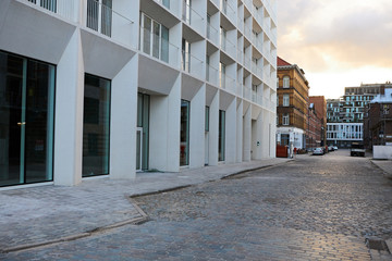 Deserted city street with modern white residential building along paved cobblestone sidewalk early in the morning with sun rising on cloudy sky in background. Clean streets of European town