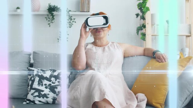 Woman seat on sofa with 3D virtual reality glasses and see color stripes. Holographic color lines around. Future concept.  Relaxing at home. 4k.
