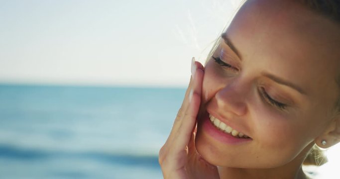 Slow motion of happy young blond hair woman is applying a sunscreen or sun tanning lotion to take care of her skin during a vacation on a beach and smiling in camera.