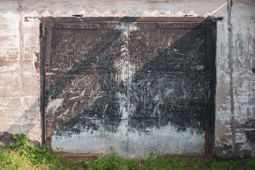 Old grunge garage door with black iron doors and iron door hinges. The gates are all scratched and scribbled. Around the plastered wall. Bottom green grass.