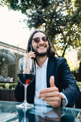 Young handsome smiling brunette bearded man joyfully talking on cellphone while resting with glass of wine in restaurant outdoor