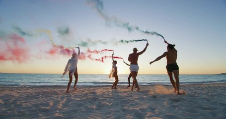 Slow motion of young multi-ethnic carefree friends in swimsuits are having fun with colorful smoke flares on a beach with a sea on a sunset.