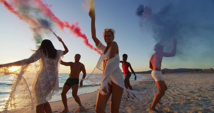 Slow motion of young multi-ethnic carefree friends in swimsuits are having fun with colorful smoke flares on a beach with a sea on a sunset.