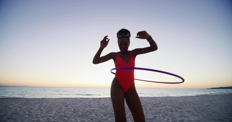Slow motion of an young african woman in bright bikini is having fun with a hula hoop on a beach with a sea on a sunset. - Powered by Adobe