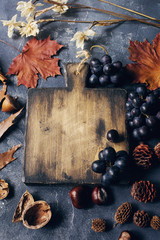 Autumn composition with cutting wooden board, marple leaves, freshly harvested ripe grapes, cones and wallnuts on dark stone table