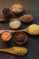 Various organic cereals and grains in wooden spoons and bowls on dark stone background