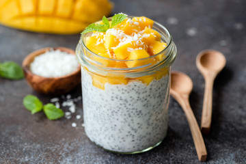 Healthy chia coconut pudding with mango in glass jar and two wooden spoons on dark background. Closeup view - 291368843