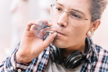 Guy with piercings and eye glasses is smoking a cigarrette.