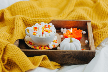 Fototapeta na wymiar A cup of coffee with marshmallow and candy corn on bed with warm plaid. Autumn beverage with candle and pumpkin on wooden tray. Hygge concept.