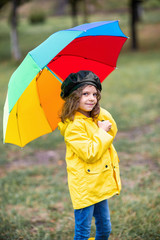 Happy funny child girl with multicolor umbrella in rubber boots at autumn park