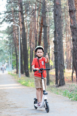 A boy in a green safety helmet rides a white scooter along a path in the forest. Bicycle helmet. Child athlete.