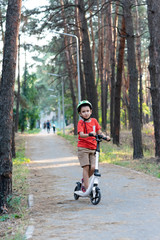 A boy in a green safety helmet rides a white scooter along a path in the forest. Bicycle helmet. Child athlete.