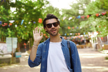 Attractive young dark haired man walking through city garden and raising hand in welcome gesture, wearing casual clothes and sunglasses