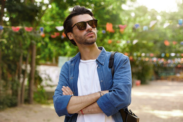 Outdoor portrait of pretty young bearded male in sunglasses posing over green park with crossed arms on his chest, looking aside with serious face