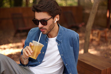 Outdoor close-up of charming dark haired male with beard wearing casual clothes and sunglasses, posing over public place, drinking juice with straw