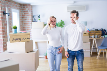 Fototapeta na wymiar Young beautiful couple standing at new home around cardboard boxes doing ok gesture with hand smiling, eye looking through fingers with happy face.