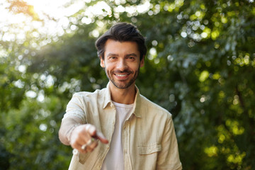 Portrait of cheerful young dark haired man looking to camera with sincere smile and raised forefinger, wearing casual clothes, posing over green park