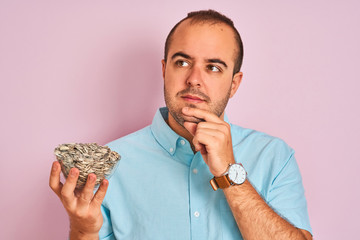 Young man holding bowl with sunflowers seeds standing over isolated pink background serious face...