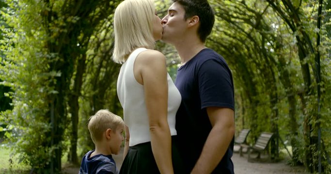 A large family walk in the Park under the plant arch: blonde mom and brunette dad kissing, mom holding her son's hand in a turtleneck and jeans, dad holding his daughter's hand in a dress.