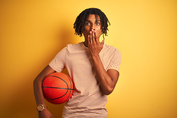 Afro sportsman with dreadlocks holding basketball ball over isolated yellow background cover mouth...