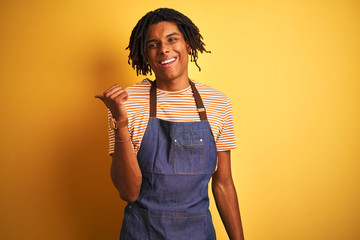 Afro american barista man with dreadlocks wearing apron over isolated yellow background smiling with happy face looking and pointing to the side with thumb up.