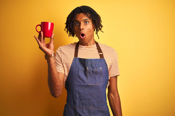 Afro barista man with dreadlocks drinking cup of coffee over isolated yellow background scared in shock with a surprise face, afraid and excited with fear expression