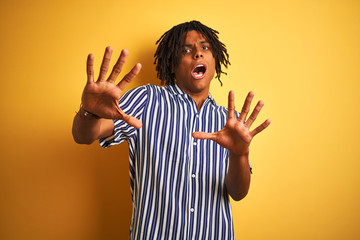 Afro man with dreadlocks wearing casual striped t-shirt over isolated yellow background afraid and terrified with fear expression stop gesture with hands, shouting in shock. Panic concept.