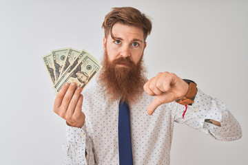 Young redhead irish businessman holding dollars standing over isolated white background with angry...