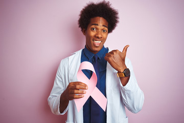 Young afro american doctor man holding cancer ribbon standing over isolated pink background pointing and showing with thumb up to the side with happy face smiling