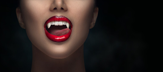 Sexy Vampire Woman's red bloody lips close-up. Vampire girl licking fangs with tongue. Fashion Glamour Halloween art design. Close up of female vampire mouth, teeth. Isolated on black background - obrazy, fototapety, plakaty