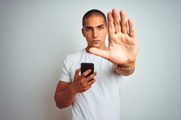 Young handsome man using smartphone over yellow isolated background with open hand doing stop sign with serious and confident expression, defense gesture