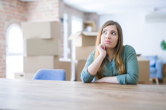 Young woman sitting on the table with cardboard boxes behind her moving to new home with hand on chin thinking about question, pensive expression. Smiling with thoughtful face. Doubt concept.
