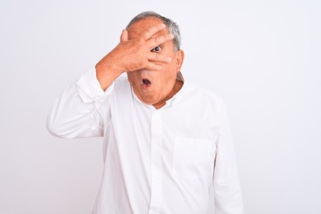 Senior grey-haired man wearing elegant shirt standing over isolated white background peeking in shock covering face and eyes with hand, looking through fingers with embarrassed expression.