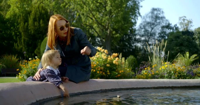 A mother with red hair and dark glasses sits with her daughter by the water of the fountain, against the background of flowerbeds, shows her somewhere with her finger.