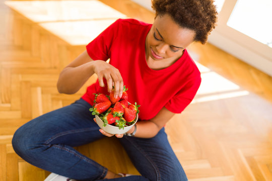 Beautiful young african woman with afro hair eating fresh strawberries sitting on the floor