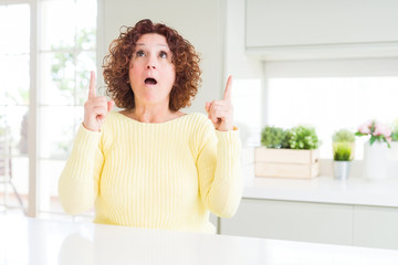 Beautiful senior woman wearing yellow sweater amazed and surprised looking up and pointing with fingers and raised arms.