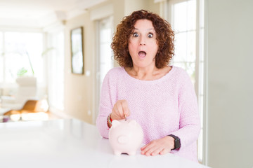 Obraz na płótnie Canvas Senior woman saving money putting a coin inside piggy bank scared in shock with a surprise face, afraid and excited with fear expression