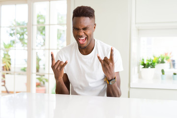 Handsome african american man on white table at home shouting with crazy expression doing rock...