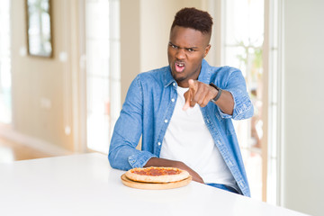 African american man eating pepperoni pizza at home pointing with finger to the camera and to you, hand sign, positive and confident gesture from the front