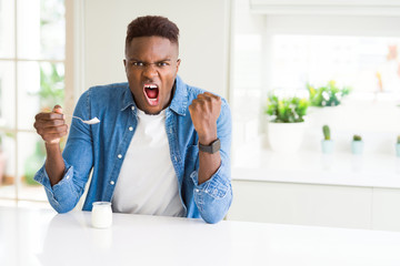 African american man eating healthy natural yogurt with a spoon annoyed and frustrated shouting with anger, crazy and yelling with raised hand, anger concept