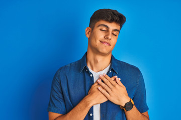 Young indian man wearing shirt standing over isolated blue background smiling with hands on chest with closed eyes and grateful gesture on face. Health concept.