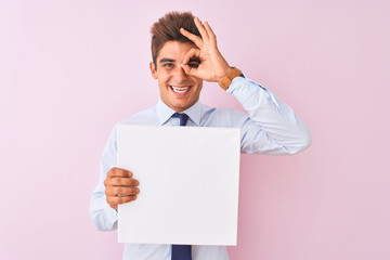 Young handsome businessman holding banner standing over isolated pink background with happy face smiling doing ok sign with hand on eye looking through fingers