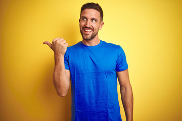 Young handsome man wearing casual blue t-shirt over yellow isolated background smiling with happy face looking and pointing to the side with thumb up.