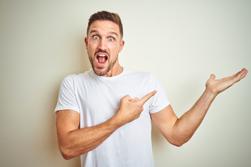 Young handsome man wearing casual white t-shirt over isolated background amazed and smiling to the camera while presenting with hand and pointing with finger.